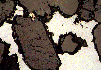 calcopirita. Pequeños granos de antimonio nativo (blanco, alta reflectancia, abajo derecha) estan atrapados junto con la bournotita. Las áreas negras son hoyos resultantes del pulido.