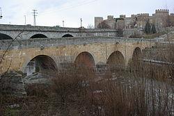 Puente romano de Ávila sobre el río Adaja, al fondo la Muralla de Ávila En todos los arcos aparece claramente la rotura precedente a la