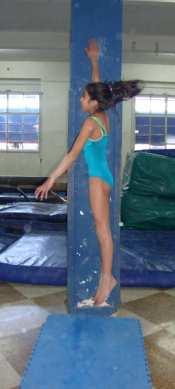 La gimnasta se coloca tiza o magnesio en su mano. se pone de costado junto a la pared, sin despegar los pies del piso, y con el brazo ectendido hacia arriba presiona la pared con su mano.